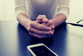 A persons hands clasped with a cell phone sitting in front of them waiting.
