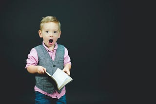 Little boy in collard shirt and vest holding a book with mouth agape in shock at what he read.