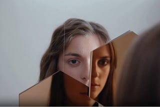 A young woman looks at herself in a broken mirror. Her reflection is split across the shards, showing different parts of her face.