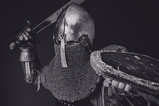 A black-and-white image of an armoured fighter (head and shoulders visible) holding up a sword in his right hand.