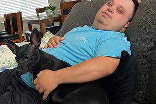 Man with Down Syndrome reclining on the couch with a little black dog leaning up against him