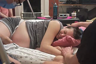 A photo of the author lying on a pile of pillow during labour. A midwife is holding a hand-held Doppler to their stomach and their partner is stroking their hair. They are wearing a t-shirt that says ‘surrogate’. They have their eyes closed and are smiling.