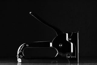 Black and silver multi-tool made for driving staples into something with more force than an ordinary office stapler. Dimly lit, the image is defined by its highlights.