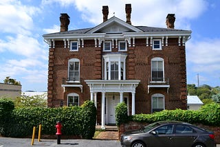 A photograph of a beautiful red brick building framed by a blue sky with a few clouds. The photo shows the ornate chimney stacks, the fancy brickwork, and the ornate white trim. The door has a small porch with white columns.