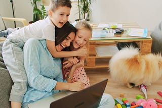 Mom on floor with laptop smiling while boy and girl hug her