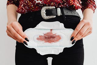 Light-skinned person in black pants with black belt and floral blouse holding a used menstrual pad horizontally in front of their pelvis.