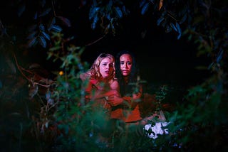 Two girls, lit in red, are seen sitting visibly scared under some lush vegetation.