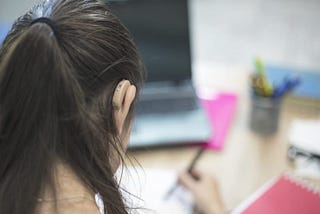 The back of a white female’s head with her brunette hair in a ponytail. We can see a skin tone behind the ear hearing aid on her right ear. She sits at a desk with a computer and paper in front of her, she is writing on the paper with a pen.