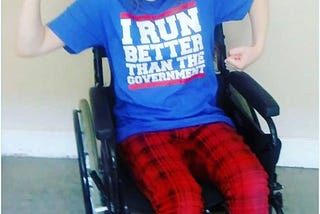 A girl in a wheelchair wearing a shirt that says, “I run better than the government”