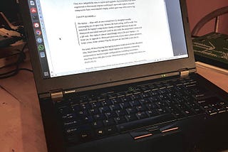 A laptop sitting on a wooden table, with a large green electronic chipboard to its right. The laptop is open and displays a Medium essay written by the author of this blog post. The laptop is thick and chunky