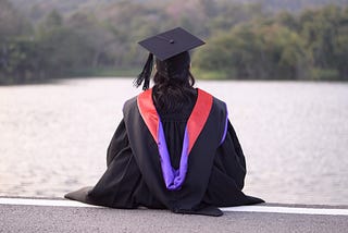 A student wearing a typical Ph.D. graduation outfit with a red and purple hood, facing away from the camera looking over a lake.