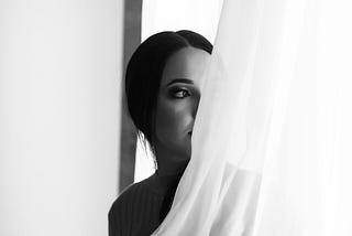 A woman standing by a window, partially veiled by a sheer curtain, holding herself in soft, introspective light.
