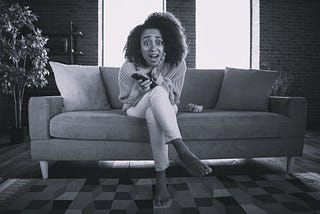 Black woman on a sofa with phone in one hand, the other hand is pressed against her face in an expression of horror and surprise.