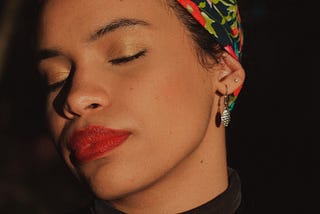 A brown skinned femme presenting person in a black turtleneck with a colorful bandana on her head and several pairs of earrings. She has gold eyeshadow and red lips and has a calm appearance with her eyes closed as the sun shines upon her.