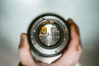 A hand holds a camera lens up against a blurred and indistinct background. In the middle of the lens the image is a sharp, well-defined picture of a city street.