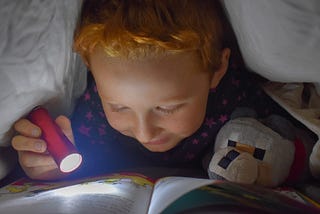 Young boy eager to learn using a flashlight and reading a book under his bed covers