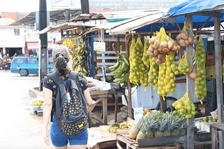 Uma mulher branca de costas, de boné e mochila, caminha de dia por barracas que vendem laranjas, abacaxis e outras frutas.