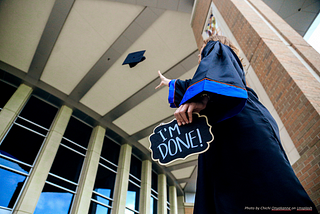 A student in a graduation gown tossing up their hat with a sign, “I’M DONE!”