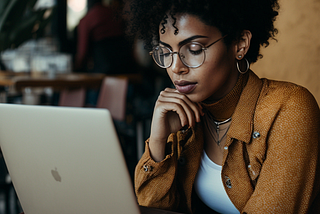 Woman with curly hair, wearing glasses sitting at a coffee shop looking at laptop.