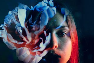 A young woman against a dark background holding a fragmented flower in front of her face and nearest to the camera. This obscures the left-hand side of her face.