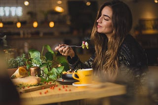 A woman with long brown hair, sitting at a wooden table at a restaurant. She is enjoying a bowl of pasta with something in a coffee mug. There is a viney plant next to her.