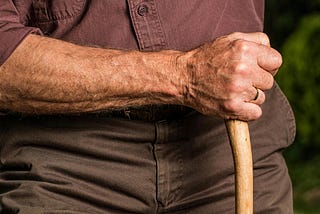 Close-up color photo of an older, but robust, man’s arm and fist gripping a walking stick.