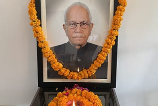 Memorial photo of an older man. Photo is decorated with marigold garland and has marigolds and a candle placed in front of it.