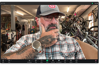 Screen capture of the Zoom user interface with the author looking at himself. He’s wearing a hat and glasses. His right hand is on his bearded chin, and his head is slightly turned, with his eyes looking directly into the camera. He’s in the thinking position. The tattoo on his hand is being primently displayed for all on Zoom to see.