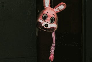 A person in a pink bunny costume with blood around its mouth leans out from a dark doorway.