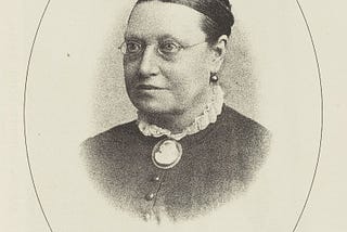 Head and shoulders photographic portrait of a Victorian woman wearing round glasses and a cameo brooch.