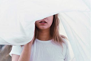 Female with brown hair wearing white top covering top half of her face with white sheet