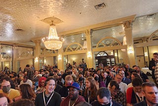 [ID: A room in the Austin Club packed with screenwriters of various genders and skin tones. The room has a white ceiling, cream colored arches, and a chandelier.]