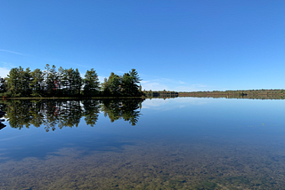 A picture of a calm and clear lake in Maine with the reflection of trees in the water.