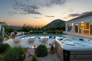 A serene outdoor setting featuring a luxurious villa with large glass doors opening to a spacious patio. The patio is equipped with modern lounge chairs around a sleek, rectangular swimming pool. Adjacent to the pool is a bubbling hot tub. The area is beautifully landscaped with diverse greenery, including ornamental grasses and purple flowering plants. In the background, a gentle hill rises under a partially clouded sky during a picturesque sunset, casting a warm glow over the entire scene.