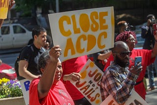 Organizers stand with red shirts, one woman has sign that says “Close ACDC” and another has a sign that says “Jobs Not Jails”