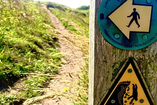 An uphill view of a winding path along a cliff edge. There are two signs in the foreground that give warning to walkers to keep to the path to avoid a dangerous fall.