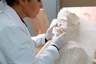 Foto von Hiba Jomaa in weißem Kittel und Handschuhen, wie sie den Zustand einer Gipsfigur betrachtet.