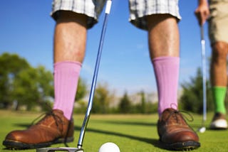 A person with bright pink socks, plaid shorts, and brown shoes about to hit a putt.