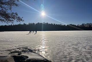 3 persons ice-skating on a frozen lake. Blue skies, the sun is shining. Frozen white lake with glimmering sun over the ice. Forests and trees in the surroundings.