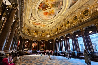 A large hall with marble pillar all around and a ceiling with an oval central painting, and rectangular ends with paintings in them. Arches embellished with gold behind an upper balcony are above the pillars.