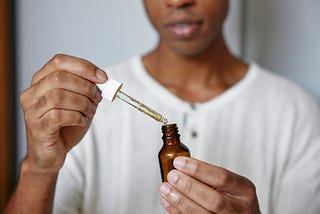 A person wearing a textured, off-white shirt is holding a small amber glass dropper bottle in one hand and a dropper filled with a light-colored liquid in the other. The dropper is being removed from or lowered into the bottle, and a droplet is visible at the tip. The person’s face is partially visible, softly focused in the background, while the hands and bottle are in clear focus in the foreground.