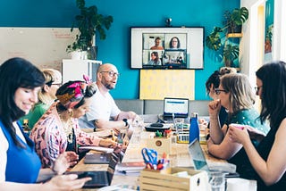 7 members of the Clearleft team sit around a large table in their studio having a meeting. A further 4 team members have joined remotely and can be seen on the TV in the background.