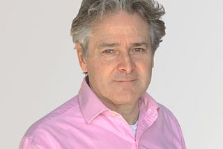 A headshot of a man with graying hair and a warm smile, wearing a pink button-down shirt and looking directly at the camera against a plain, light background.