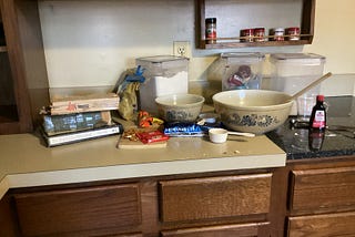 Kitchen counter covered with baking materials and a stack of old cookbooks, illustrating reading to learn to cook rather than being taught by another person.