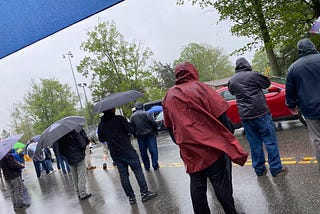 A line of people standing in rain, along the entrance to a manufacturing plant.