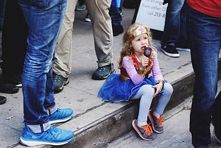 A small girl dressed as Wonder Woman, sitting on a curb, surrounded by adults (legs only), and eating ice cream.