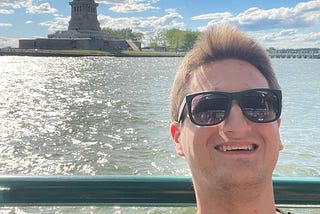 Man wearing sunglasses on boat with Statue of Liberty in background.