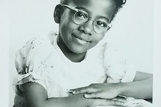 Little black girl wearing glasses, seated at desk, smiling