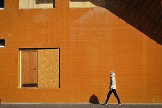 Pessoa caminhando sozinha na frente de uma parede ocre, com uma porta meio tapada na frente.