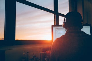 man sitting facing back in front of laptop next to window as the sun sets